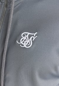 Veste zippée grise avec une texture lisse, arborant un logo brodé blanc sur le côté gauche. Accentuée par un cordon de serrage tressé.