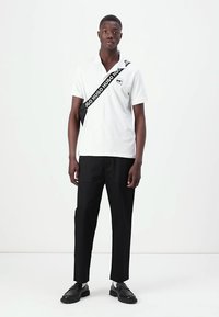 Biely polo tričko s golierom, na ktorom sa nachádza logo a čierny pás s textom "HUGO" naprieč hrudníkom. Čierne nohavice a čierne topánky dopĺňajú outfit.