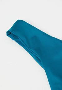 Gros plan du tissu du bas de bikini turquoise avec une ceinture pliée et une texture lisse sur fond blanc.