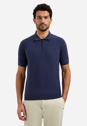 Man met donker krullend haar, gekleed in een marineblauw poloshirt met korte mouwen en ritssluiting, en lichtbeige broek, staand met één hand in de zak.
