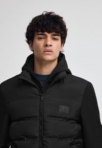 Svart puffjacka med dragkedja, texturerad front och släta ärmar. Har en huva och en liten präglad logotyp på bröstet.