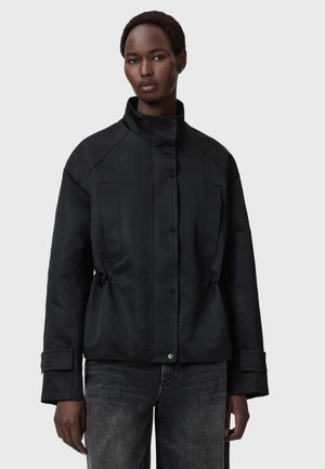 Tummaihoinen henkilö, joka pukeutunut mustaan korkeakauluksiseen takkiin, jossa on nappisuljenta edessä, ja mustiin farkkuihin, seisomassa yksiväristä vaaleaa taustaa vasten.