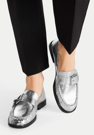 Srebrne mokasyny o teksturowanym wykończeniu, z ozdobnym metalowym detalem i płaską czarną podeszwą, noszone z czarnymi zwężanymi spodniami.