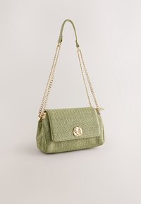 Sac à main tissé vert avec une bandoulière chaîne en or et un fermoir en or irrégulier sur le rabat avant, présenté sur un fond clair uni.