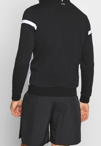 Felpa nera con dettagli bianchi, polsini e vita a costine, con colletto alto. Abbinata a pantaloni corti neri, mano visibile.