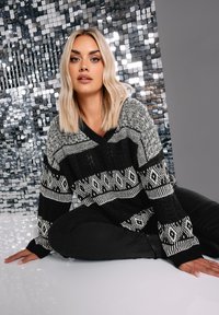 Schwarzer und weißer gemusterter Pullover mit V-Ausschnitt, der geometrische Designs, eine lockere Passform und weite Ärmel aufweist. Modell sitzt vor einem spiegelnden Hintergrund.