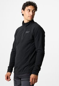 Schwarzer Fleece-Pullover mit Reißverschlusskragen, weichem Textil und seitlichen Nähten. Logo auf der Brust. Das Modell trägt dunkle Hosen.