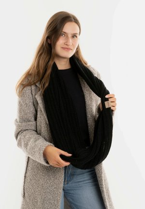 Zwarte gebreide infinity sjaal vastgehouden door een persoon, met een gestructureerde stof en een klein merklabel. Geddragen met een lichtgrijze jas en denim.