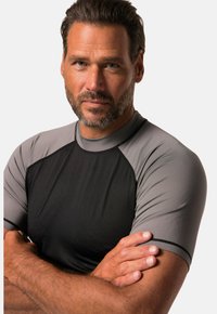 Ciemnoszara i jasnoszara koszulka sportowa z krótkimi rękawami, gładka w dotyku, z kontrastującymi przeszyciami. Zbliżenie na pozę z założonymi rękami.