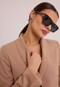 Czarne trójkątne okulary przeciwsłoneczne z grubymi oprawkami; modelka ubrana w beżową marynarkę dopasowaną z teksturowanej tkaniny; duże okrągłe srebrne kolczyki.