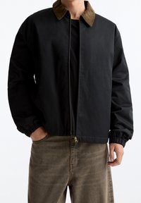 Zwarte zip-up jack met een bruine corduroy kraag gedragen over een zwart shirt, gecombineerd met losse bruine broek, handen in de zakken.