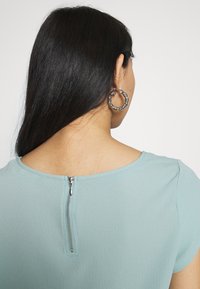 Ljusblå kortärmad topp med rund halsringning, med en synlig dragkedja i ryggen och strukturerad tyg. Stora vridna silverörhängen.