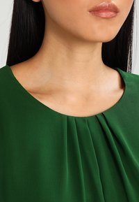 Primo piano del collo e delle spalle di una persona che indossa una blusa verde con pieghe, caratterizzata da una pelle liscia e capelli scuri e lisci.