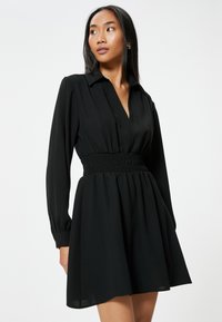 Schwarzes Kleid mit gerafftem Taillenbereich, langen Ärmeln und einem Kragen. Der Stoff ist glatt und der fließende Rock endet oberhalb des Knies.