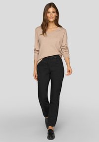 Beiger Pullover mit V-Ausschnitt, weiche Textur; schwarze gerade geschnittene Hose, glatter Stoff; schwarze Loafers; Modell steht vor einem grauen Hintergrund.