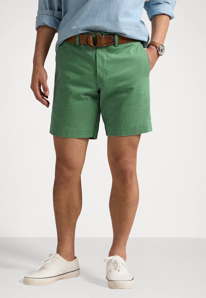 Zaļas kokvilnas šorti ar plakanu priekšpusi un sānu kabatām, papildināti ar brūnu ādas jostu un baltām auduma sporta kurpēm.