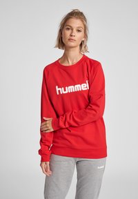 Felpa rossa con logo bianco "hummel", caratterizzata da un colletto rotondo e polsini a coste. Abbinata a pantaloni della tuta grigi, per un look informale.