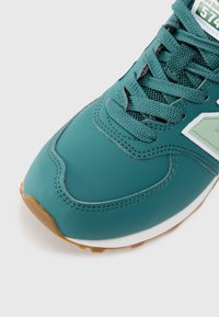 Teal värvi New Balance 574 sneakers, mille esiosa on tekstureeritud paeltega, õmmeldud overlay-dega, valge keskmise tallaga ja kummist väljatallaga valgel taustal.