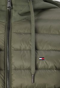 Grøn pufferjakke med lynlås foran, med en struktureret overflade, ribstrikkede snore og et lille emblem i rød, hvid og blå.