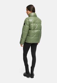 Pufferjakke i skinnende olivengrøn med høj krave, vandret syninger og et sort logo-flag. Båret over sorte skinny jeans og støvler.