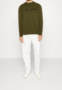 Felpa verde oliva con testo e logo "TOMMY HILFIGER" neri, abbinata a chinos bianchi e sneakers bianche su uno sfondo neutro.