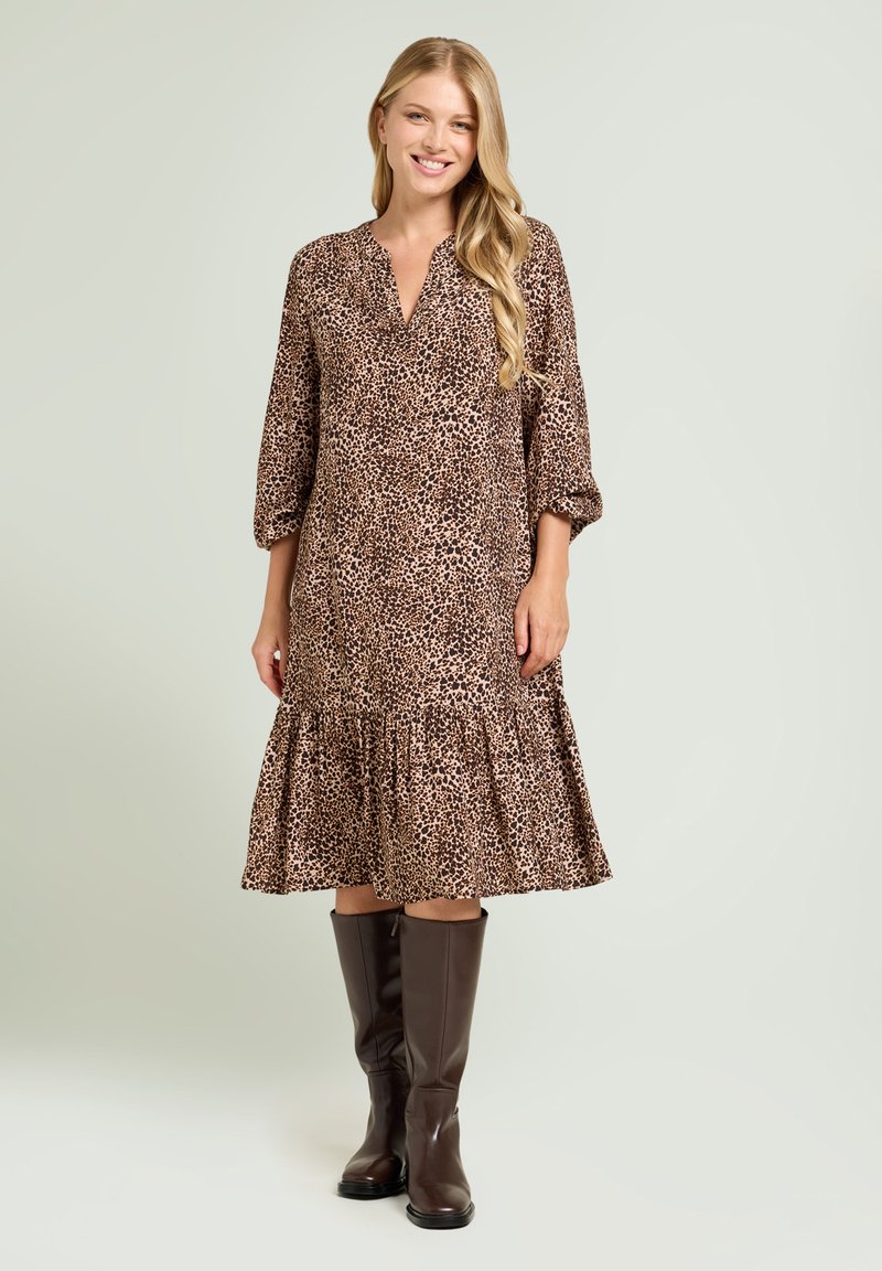 Leopardenmuster Kleid mit langen Ärmeln, V-Ausschnitt und gestuftem Volant-Saum. Kombiniert mit kniehohen braunen Lederstiefeln. Glatte Stoffstruktur.