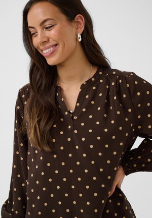 Femme aux longs cheveux foncés souriante, portant des boucles d'oreilles anneaux en argent et un chemisier marron à pois beiges avec une petite ouverture en V.