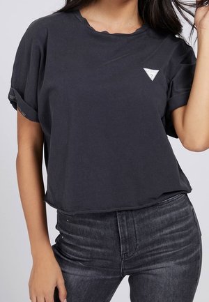 Kobieta w ciemnoszarej krótkiej koszulce z krótkim rękawem i małym białym trójkątnym logo oraz ciemnoszarych, wysokich dżinsach.