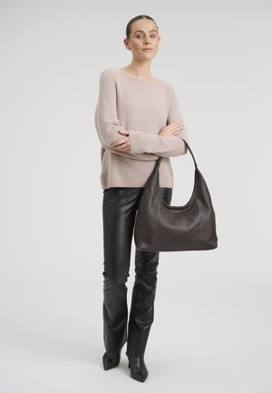Žena stoji s prekriženim rukama, nosi svijetloružičasti džemper, crne kožne hlače, crne šiljate čizme i drži veliku tamnocrnu kožnu torbu.