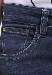 Los vaqueros de mezclilla azul oscuro tienen una cintura alta, un bolsillo redondeado en la parte delantera y un acento de estrella dorada. La tela tiene una textura suave con costuras en contraste.