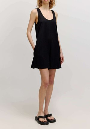 Schwarzes ärmelloses Kleid mit rundem Ausschnitt, fließendem Stoff, seitlichen Taschen und lockerer Passform, kombiniert mit schwarzen flachen Sandalen.