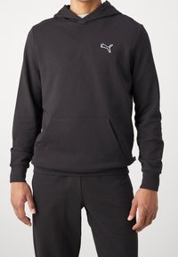 Sudadera negra con capucha tipo pulóver y bolsillo frontal, con un pequeño logo blanco de Puma en el pecho. Fabricada con tela suave y capucha con cordón ajustable.
