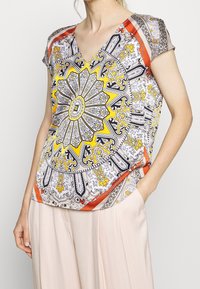 Blouse à manches courtes présentant un motif circulaire vibrant en jaune, noir et orange sur un fond clair, avec un décolleté en V.
