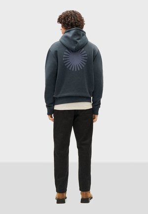 Donkerblauwe hoodie met een ronde graphic op de achterkant, textuurstof en een relaxte pasvorm. Gecombineerd met zwarte broek en casual schoeisel.