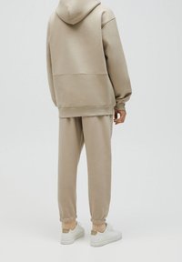 Sweat à capuche beige avec une grande capuche et une poche kangourou, associé à un pantalon de survêtement assorti avec poignets resserrés. Tissu lisse et coupe décontractée. Baskets blanches.