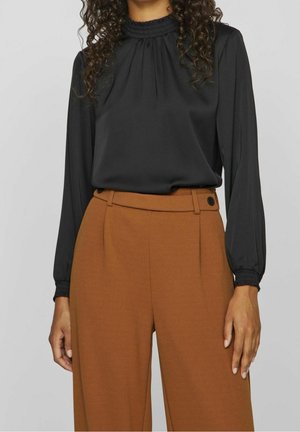 Kvinde iført sort langærmet bluse med rynkede manchetter og høj hals, kombineret med højtaljede brune bukser med vide ben.