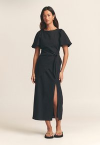 Czarna sukienka do kolan z krótkimi bufiastymi rękawami, kopertowym fasonem i przednim rozcięciem. Noszona z płaskimi, jasnobrązowymi sandałami. Gładka faktura materiału.