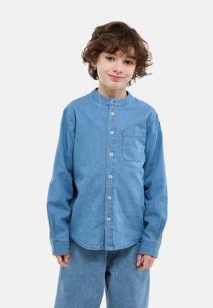 Enfant aux cheveux bouclés bruns portant une chemise boutonnée en denim bleu et un jean assorti, debout devant un fond uni.