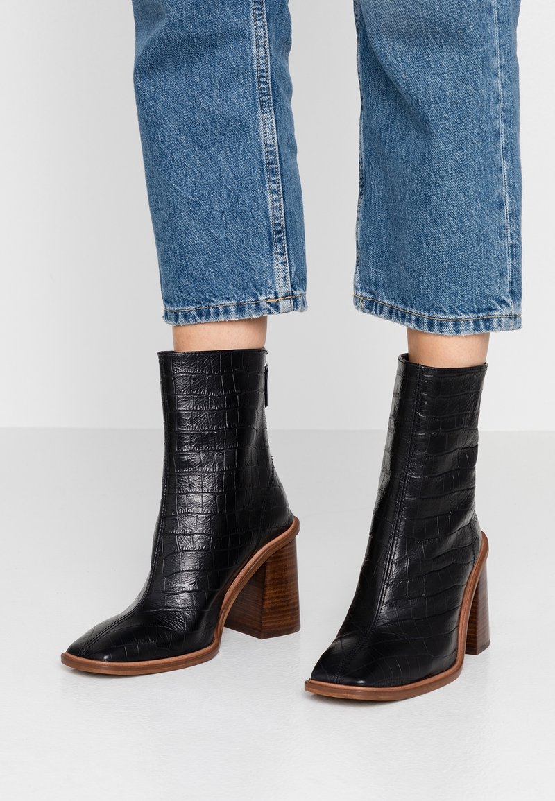 Bottes de cheville noires en relief crocodile avec un bout pointu et un talon en bois épais, assorties à un jean en denim bleu raccourci.