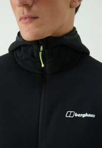 Crna zip-up jakna s kapuljačom, s blistavim zelenim povlačenjem za patentni zatvarač i bijelim logom na prsima. Glatka tekstura i uski kroj.