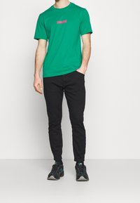 Zelené bavlněné tričko s růžovým grafickým logem, v kombinaci s černými slim-fit kalhotami a černými teniskami s tyrkysovými akcenty.