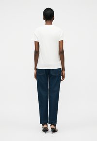 Mujer de cabello corto de espaldas, vestida con una camiseta blanca de manga corta, jeans azul oscuro y sandalias negras de tacón alto.