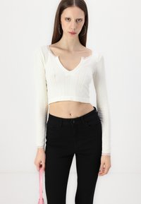 Biela krátka top s háčkovým vzorom a výstrihom do V, s krajkovým lemovaním; kombinovaná s čiernymi vysokými skinny džínsami a ružovou kabelkou.