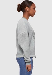 Grå sweatshirt med långa ärmar, rund halsringning och dragkedjor på sidorna. Har grafisk tryck på framsidan och ribbade muddar. Tillsammans med blå jeans.