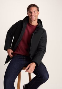 Smiling man sitting on wooden stool wearing black hooded jacket, dark blue jeans, and maroon sweatshirt against plain background.