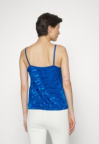 Top azul sem mangas com um padrão ondulado, feito de tecido leve, apresenta alças finas e um corte solto, combinado com calças brancas.