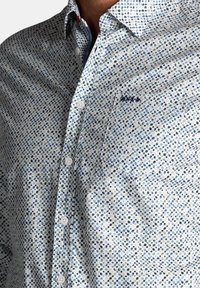 Overhemd van lichte stof met een witte basis en een blauw-groene stippenpatroon. Inclusief een borstzak en geborduurd logo.