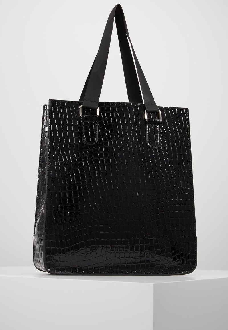 Sac cabas noir brillant avec texture peau de crocodile et doubles anses plates exposé sur un socle blanc sur fond gris.