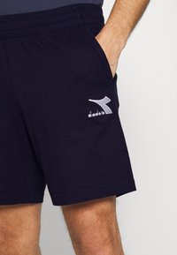 Shorts de sport bleu marine en coton avec une taille élastique et des poches latérales. Présente un logo brodé blanc sur le côté gauche.