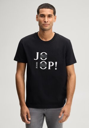 Mann trägt ein schwarzes T-Shirt mit weißem Text "JOOP!" und "JEANS" und steht vor einem schlichten hellen Hintergrund.